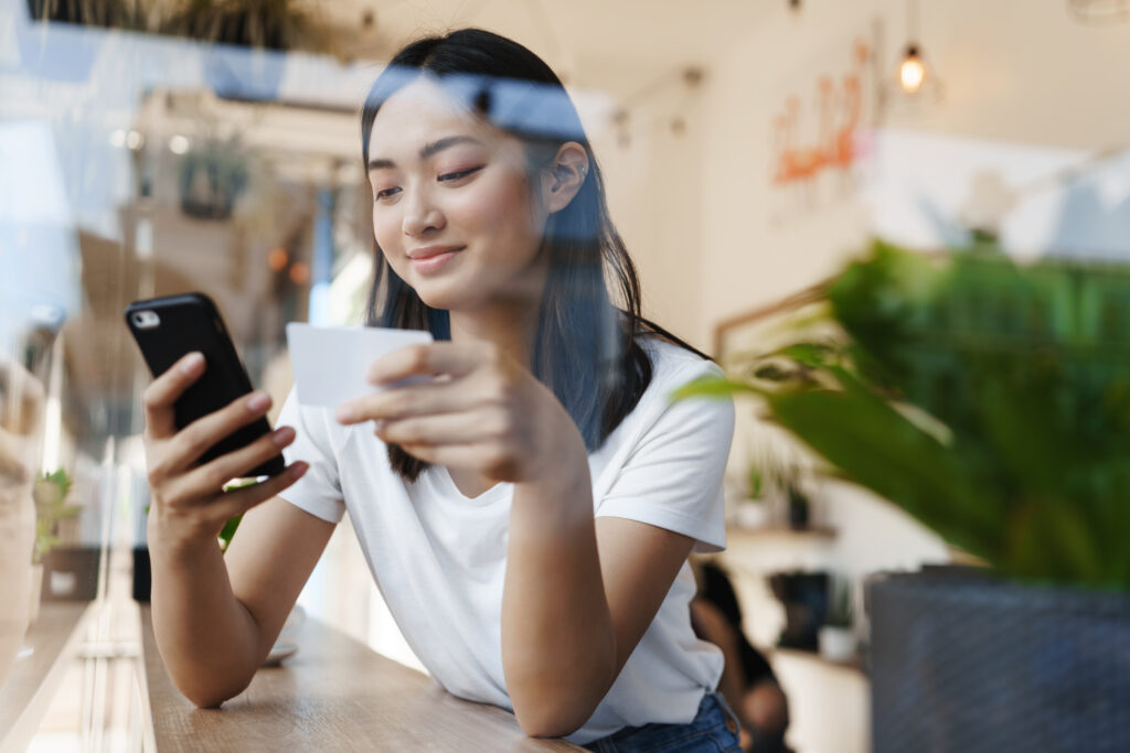Stylish asian girl sitting in cafe near window, paying for online shopping with credit card, transfer money for coffee with smartphone, casually sitting in restaurant on summer day.